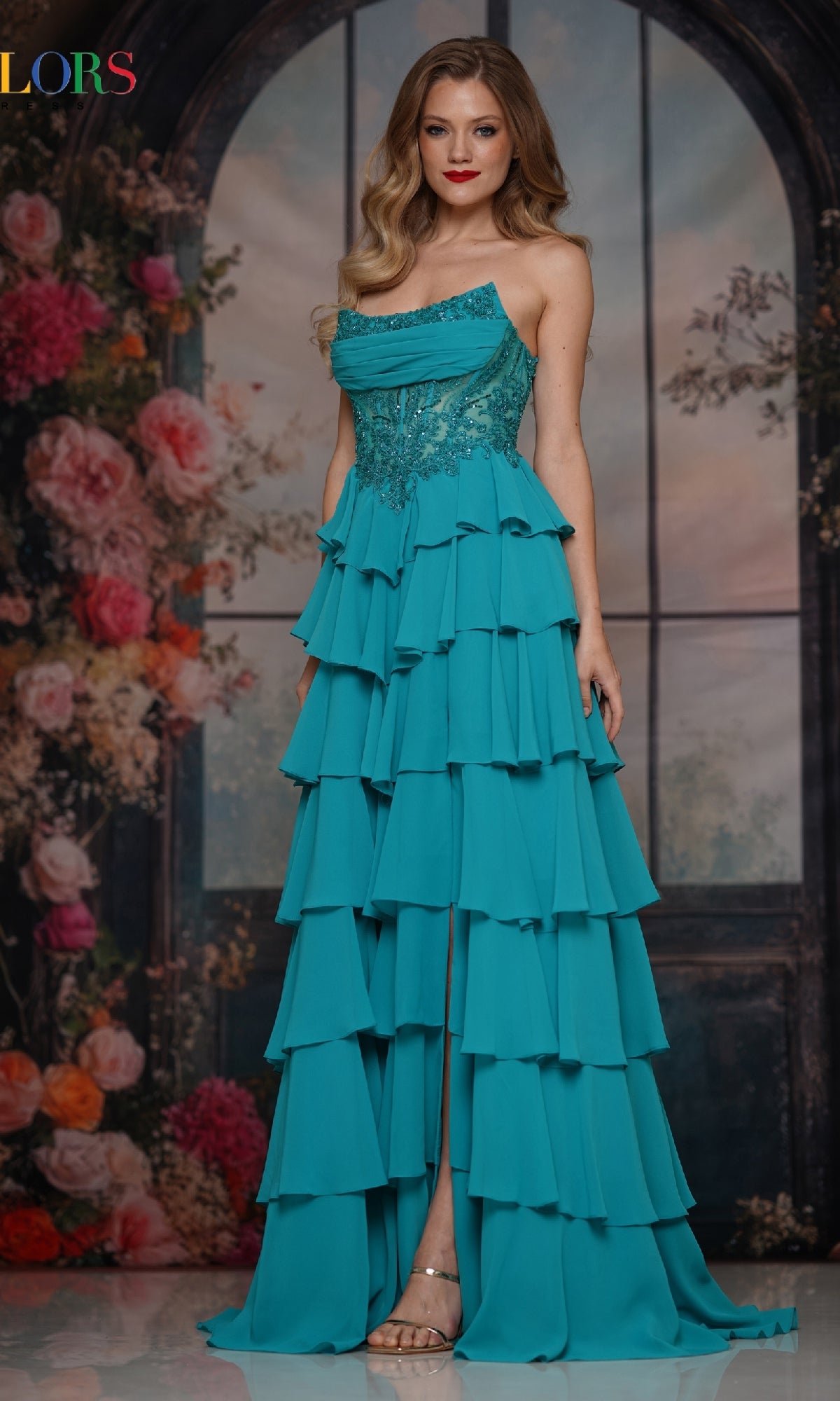 A woman in the Colors Dress 3934, a strapless long ruffled prom dress, stands indoors before a floral backdrop with loose waves and open-toed heels completing her formal look.